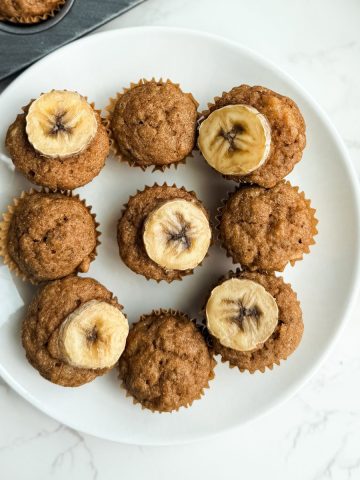Mini banana bread muffins topped with fresh banana slices arranged on a white plate, with a whole banana beside them